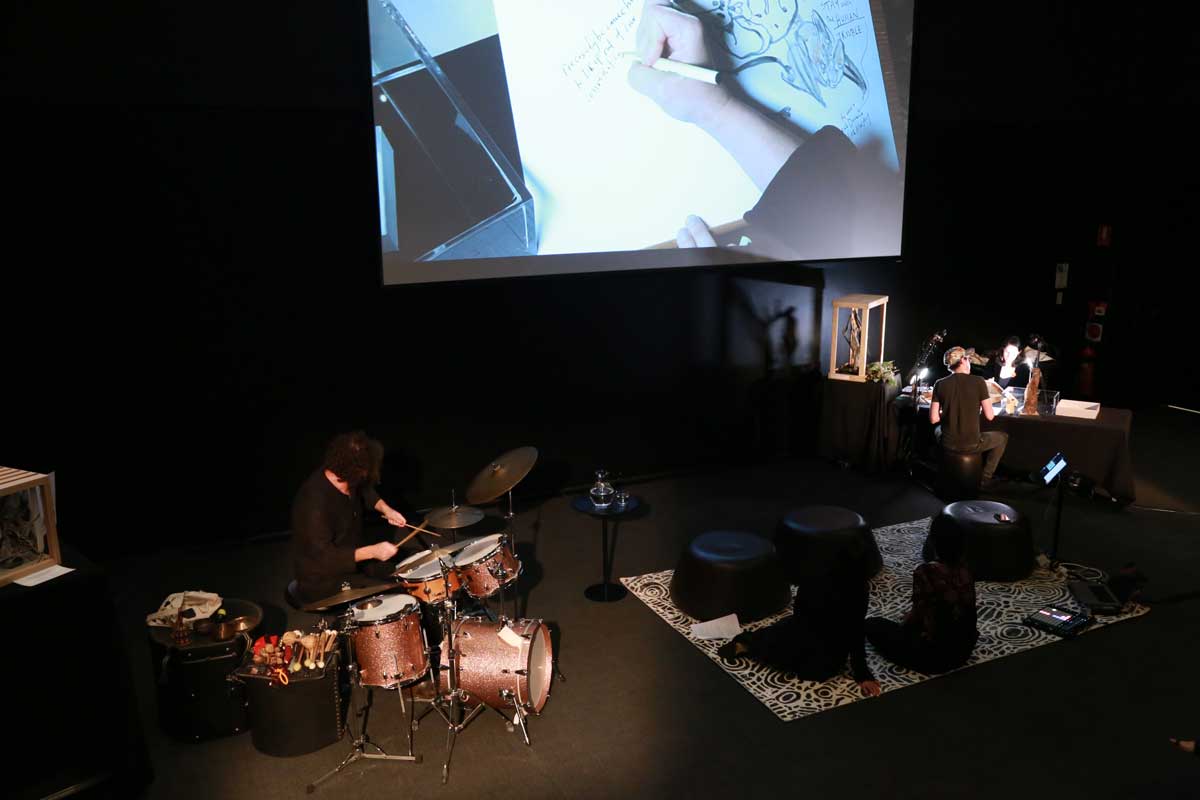 A drummer plays in front of a screen projecting hands drawing, a man and a woman are at a table nearby drawing