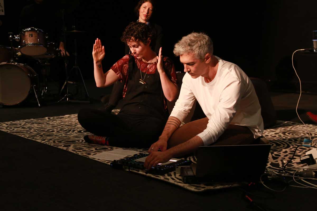 A woman and a man sit cross legged on the floor, the woman speaks while the man mixes music on an audio deck