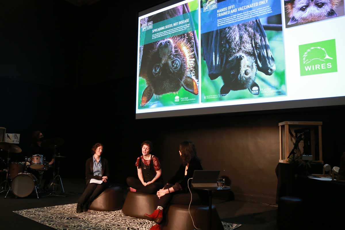 Three women in conversation, seated at the front of a theatre, a screen with images of bats is behind them.